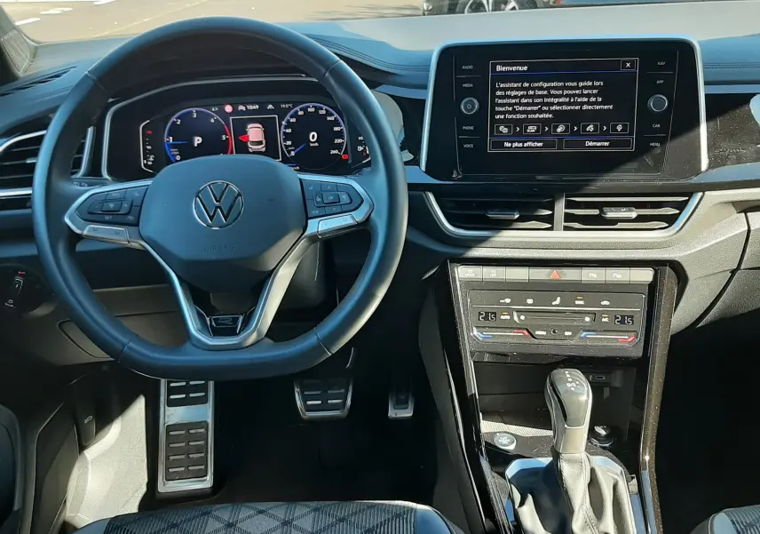Vue intérieure du tableau de bord et volant du Volkswagen T-Roc gris Indium 2024, avec écran tactile et levier de boîte automatique.