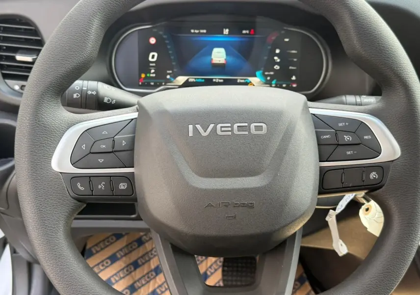 Vue rapprochée du volant noir Iveco avec commandes intégrées et tableau de bord numérique en arrière-plan.