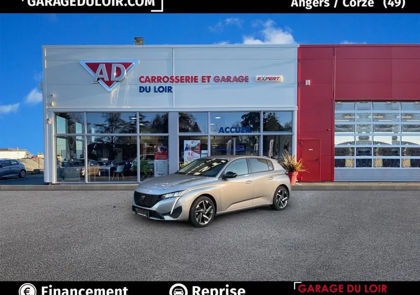 Peugeot 308 Gris Artense vue de 3/4 avant garée devant un garage avec calandre noire et jantes alliage 17 pouces.