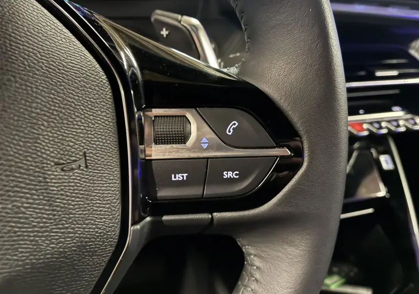 Gros plan sur les commandes gauche du volant noir du Peugeot 2008 1.2 Hybrid 136 Allure, avec boutons rétroéclairés.