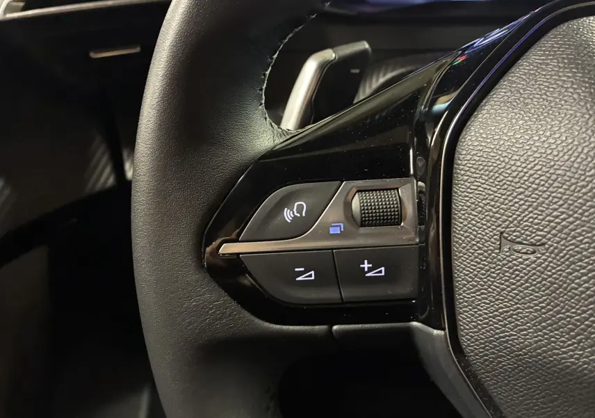 Gros plan sur les commandes gauche du volant noir du Peugeot 2008 1.2 Hybrid 136 bleu, avec molette et boutons lumineux.