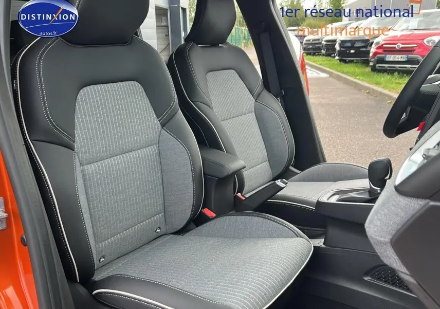 Vue intérieure côté conducteur de la Renault Clio V orange Valencia, sièges sport gris avec surpiqûres blanches et accoudoir central.