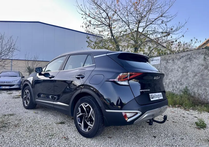 Vue 3/4 arrière droit du Kia Sportage noir 2023 avec attelage et jantes alliage distinctives sur un sol gravillonné.