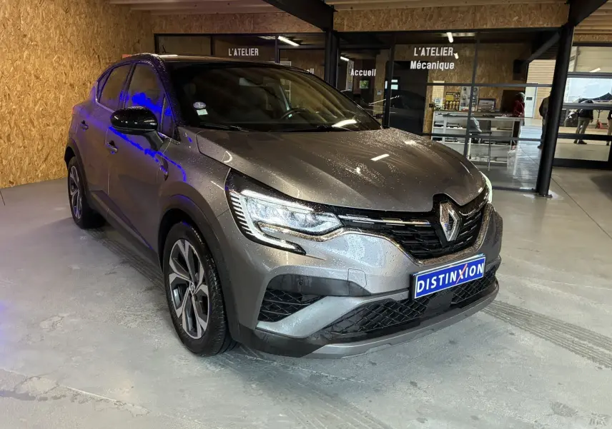 Renault Captur gris 3/4 avant droit avec phares allumés, garé en intérieur devant un atelier mécanique.