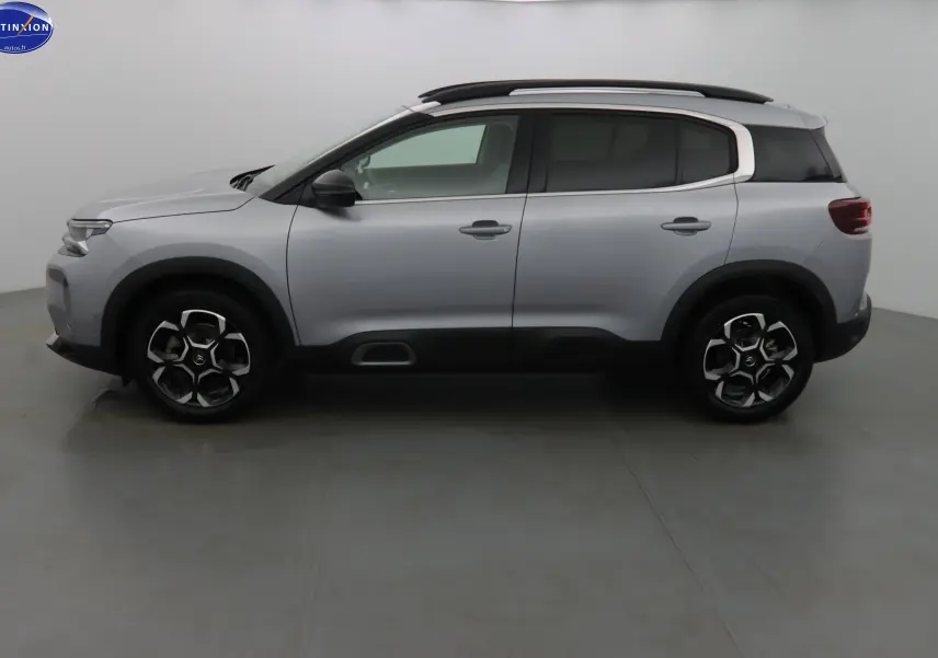 Profil latéral gauche d'un Citroën C5 Aircross gris Artense métal avec barres de toit et protections noires sur les passages de roues.