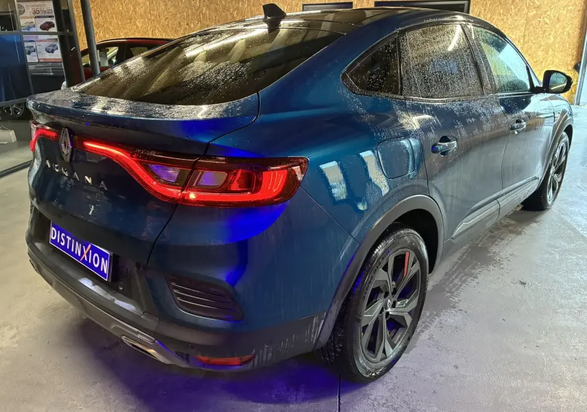 Vue 3/4 arrière droite d'une Renault Arkana bleu 2021 avec feux arrière allumés et jantes noires distinctives.