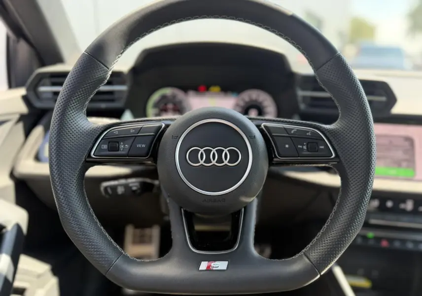 Gros plan sur le volant sport en cuir perforé avec logo Audi et badge S, intérieur moderne et lumineux.