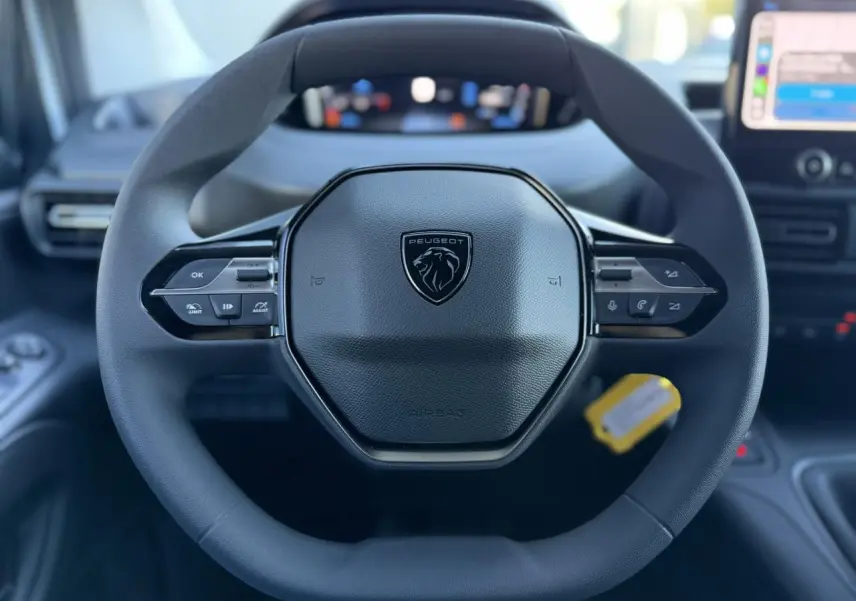 Vue rapprochée du volant noir multifonctions du Peugeot Partner 2026 avec combiné numérique flou en arrière-plan.
