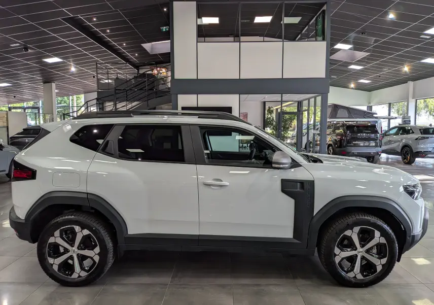 Profil droit du Dacia Duster Hybrid-G blanc glacier 2026 en showroom, avec jantes bicolores et protections latérales noires.