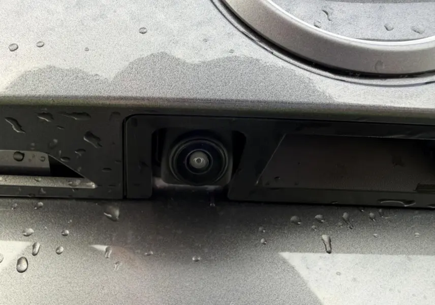 Gros plan sur la caméra de recul intégrée à la plaque arrière du Nissan Juke II noir métal, avec gouttes d'eau visibles.