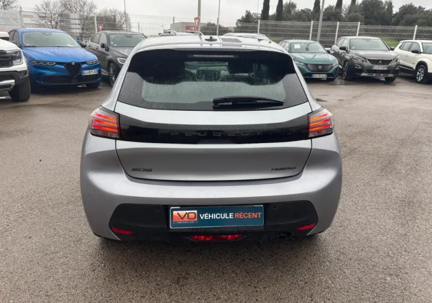 Vue arrière d'une Peugeot 208 Hybrid gris 2025 avec feux arrière allumés sur parking humide.