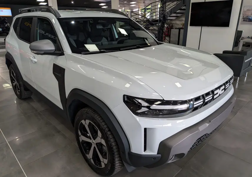 Vue 3/4 avant droite du Dacia Duster Hybrid-G blanc Glacier avec détails noirs contrastants et jantes bi-ton en showroom.