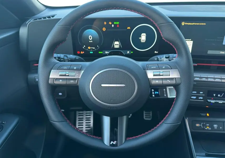 Vue intérieure centrée sur le volant cuir noir avec surpiqûres rouges du Hyundai Kona 2025, tableau de bord numérique visible.