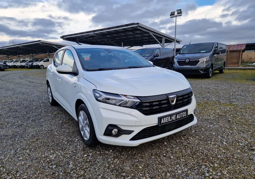 Dacia Sandero blanc vue 3/4 avant droit sur parking gravillonné avec plusieurs véhicules en arrière-plan.