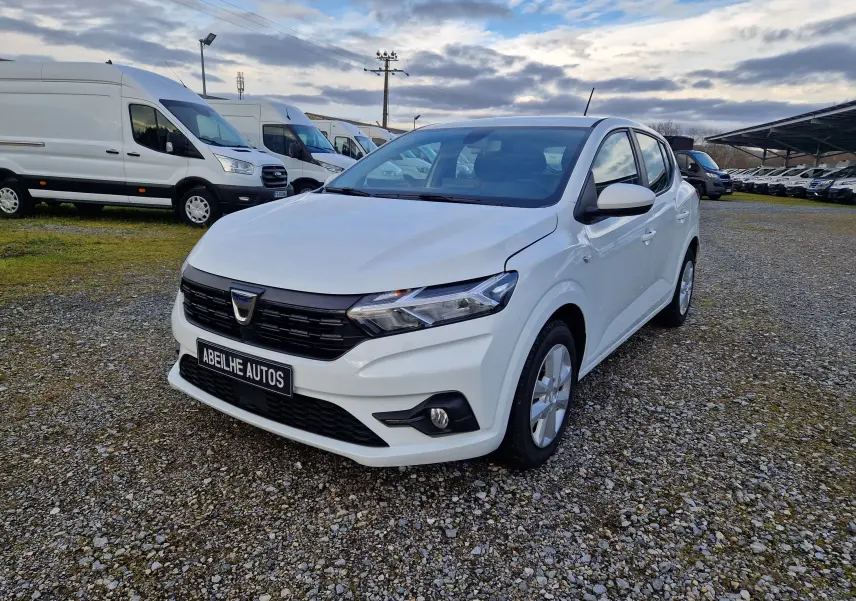 Dacia Sandero blanc vue 3/4 avant droit sur parking gravillonné avec plusieurs fourgons blancs en arrière-plan.