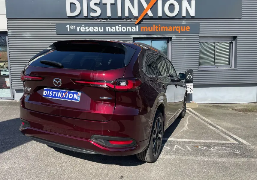 Vue 3/4 arrière droite du Mazda CX-80 hybride rechargeable en rouge Artisan, stationné devant un bâtiment Distinxion.