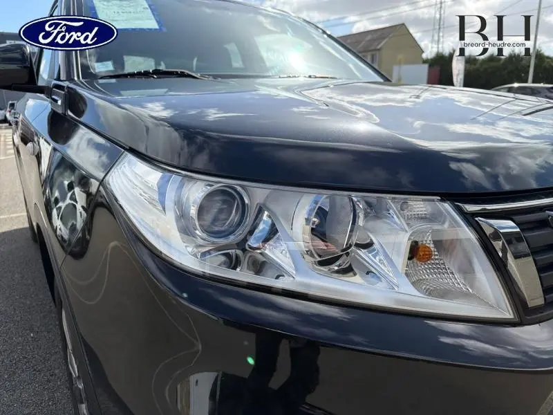 Gros plan sur le phare avant gauche du Suzuki Vitara Cosmic Black Pearl métallisé, reflétant le ciel et l'environnement.