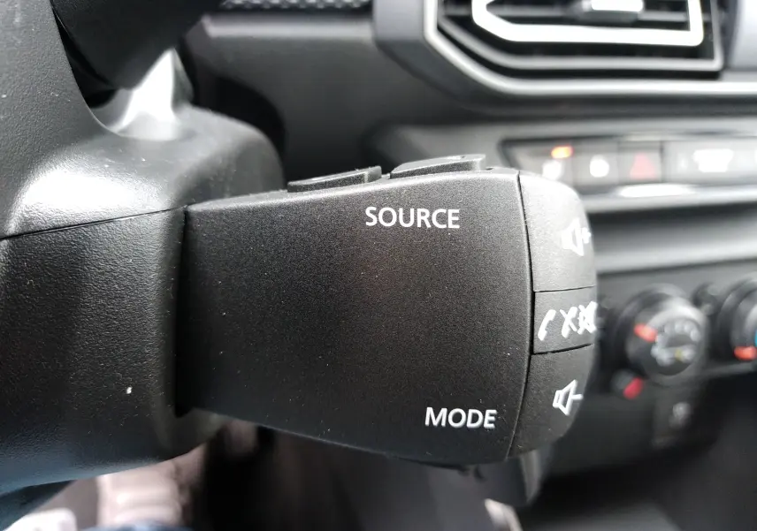 Gros plan sur la commande audio au volant du Dacia Sandero 2025, intérieur noir avec boutons source et mode visibles.