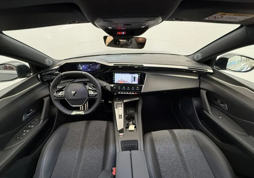 Vue intérieure avant de la Peugeot 308 Hybrid 136 e-DCS6 GT 2024, tableau de bord et sièges noirs avec volant cuir multifonctions.