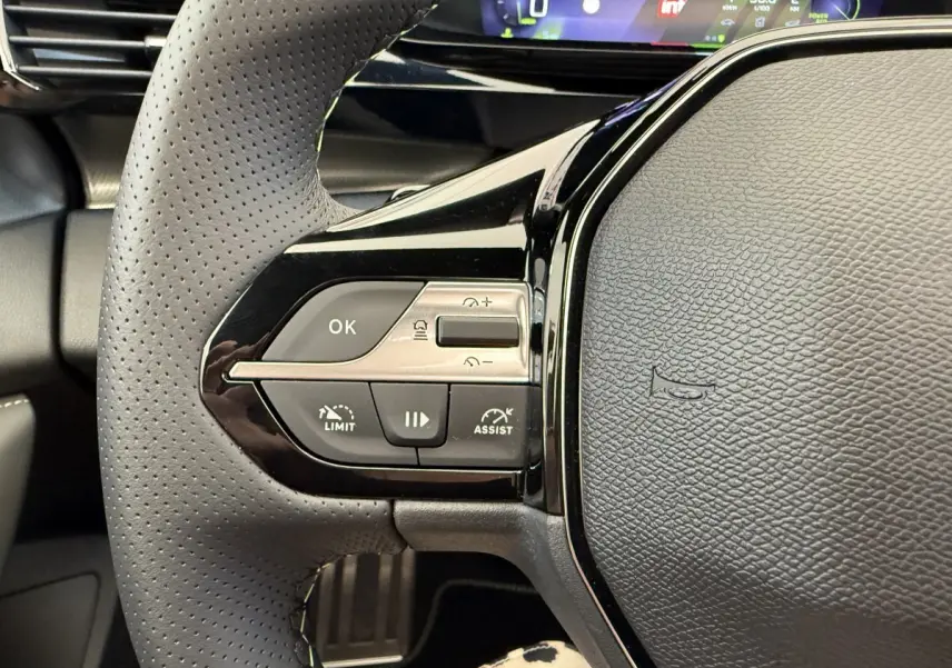 Gros plan sur le volant cuir perforé gris foncé de la Peugeot 308 Hybrid 136 e-DCS6 GT avec commandes multifonctions et tableau de bord numérique.