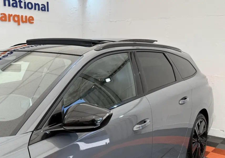 Vue côté gauche avant d'une Peugeot 308 Hybrid 136 e-DCS6 GT gris foncé avec rétroviseur noir brillant et barres de toit.