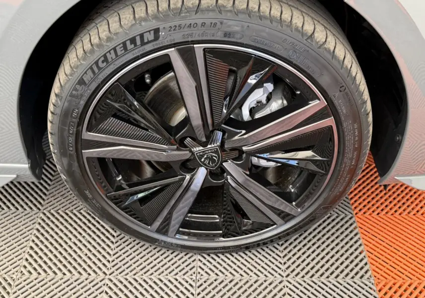 Gros plan sur la roue avant droite noire brillante d'une Peugeot 308 Hybrid 136 e-DCS6 GT gris foncé, avec logo lion central.