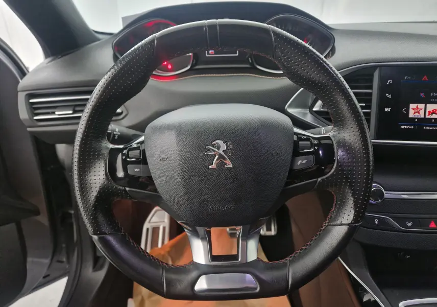 Vue rapprochée du volant en cuir perforé noir avec logo Peugeot, tableau de bord et écran tactile visibles en arrière-plan.