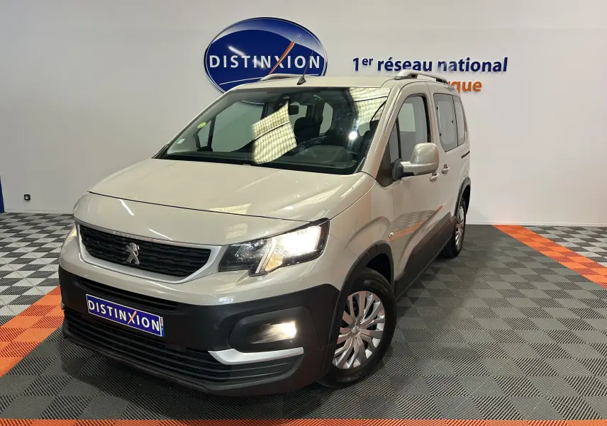 Peugeot Rifter gris sable en 3/4 avant droit, avec attelage et phares allumés dans un showroom.