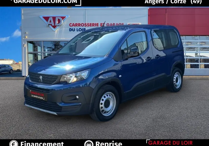 Vue 3/4 avant droite d'un Peugeot Rifter bleu garé devant un garage avec jantes tôles et rétroviseurs noirs vernies.