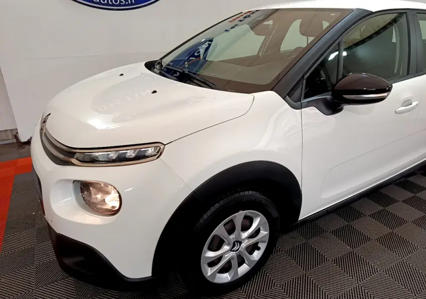 Vue 3/4 avant droit d'une Citroën C3 blanche avec phares allumés et rétroviseurs noirs contrastants.