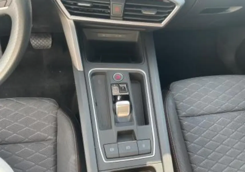 Vue rapprochée de la console centrale de la SEAT LEON IV blanche, avec levier de boîte auto et sièges sport noirs à surpiqûres rouges.