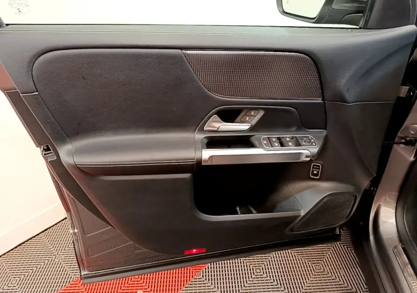 Vue intérieure côté gauche de la porte avant d’un Mercedes GLB 200 d Business Line gris foncé, avec commandes électriques et poignée argentée.