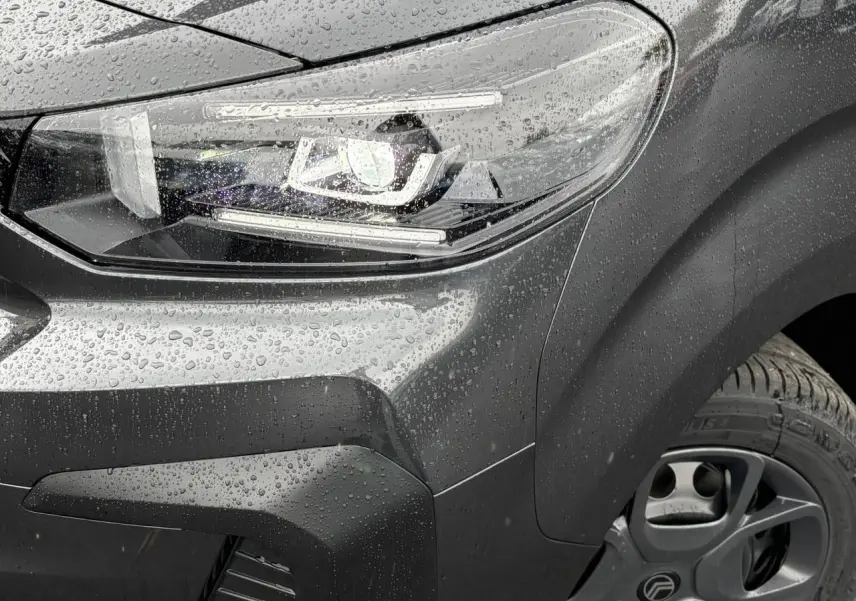 Gros plan sur le phare avant droit et la roue d'un Citroën Jumpy gris titane sous la pluie, avec détails LED visibles.