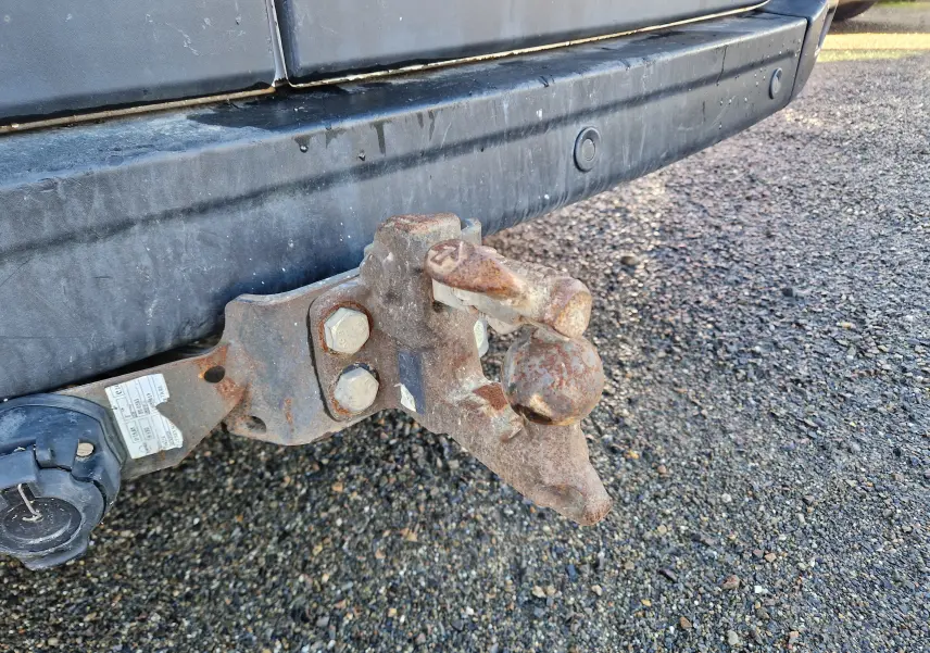 Gros plan sur l'attelage rouillé fixé au pare-chocs arrière noir d'un Renault Master blanc, vue latérale basse.