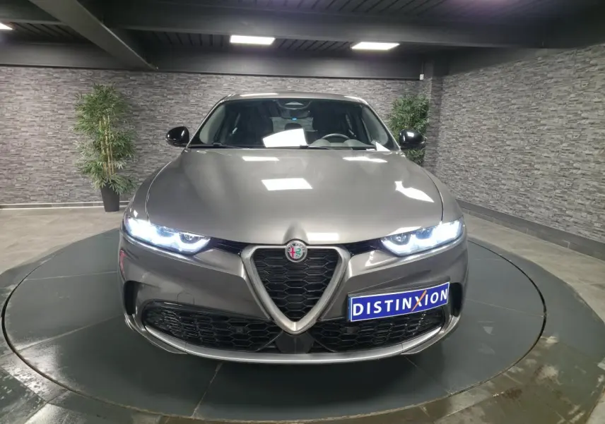Vue avant d'une Alfa Romeo Tonale 1.3 PHEV gris métallisé avec phares LED allumés en intérieur showroom.
