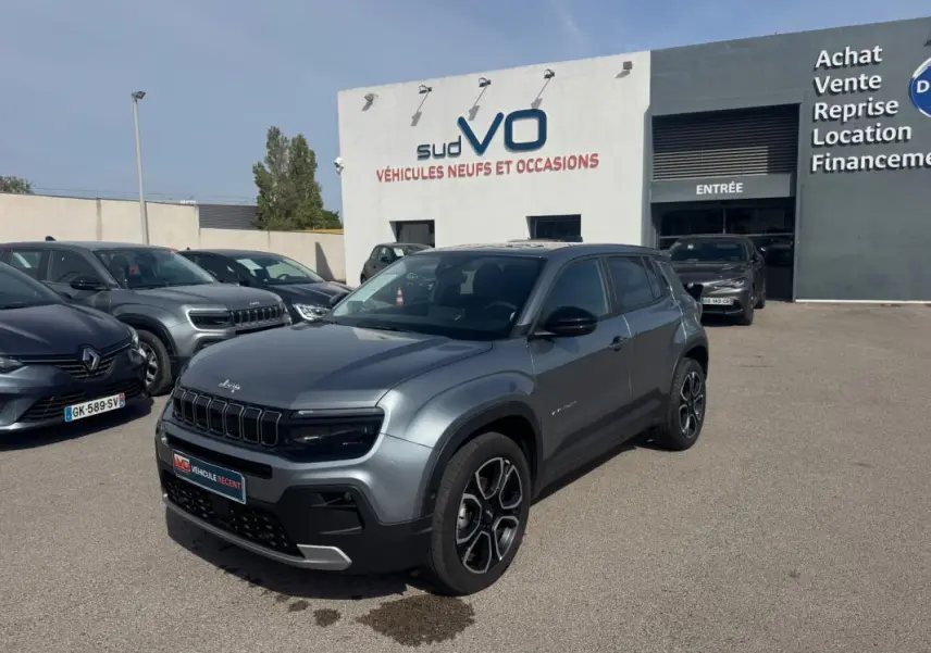 Jeep Avenger gris foncé en vue 3/4 avant droit, avec calandre noire et jantes distinctives, devant un concessionnaire.