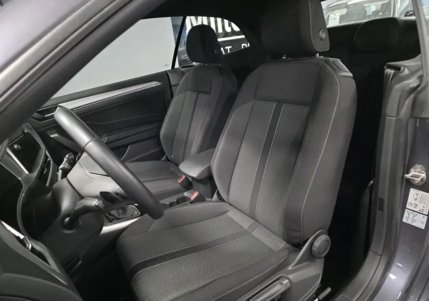 Vue intérieure côté conducteur du Volkswagen T-Roc 2024 gris, sièges tissu noir avec accoudoir central et volant multifonction.