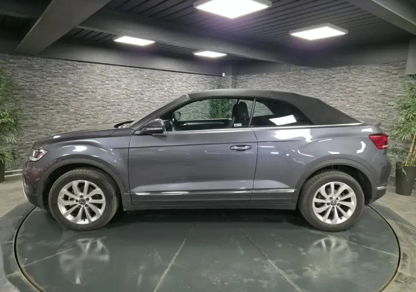 Volkswagen T-Roc Style gris métallisé vue de profil côté gauche avec capote textile noire en intérieur showroom.