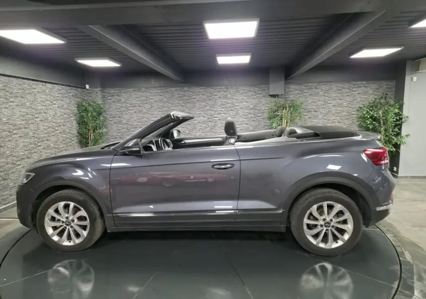 Profil droit d'un Volkswagen T-Roc cabriolet gris métallisé 2024 avec capote noire repliée en intérieur showroom.
