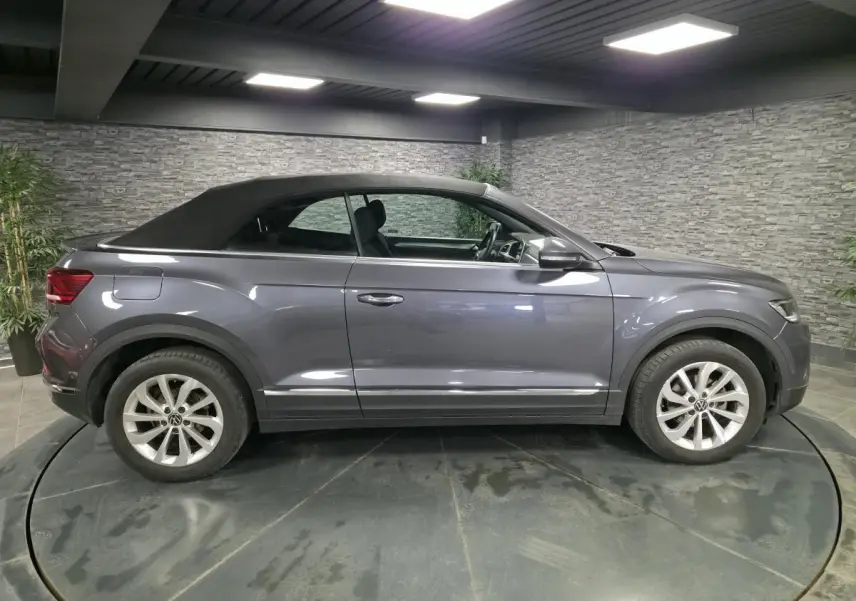 Volkswagen T-Roc gris métallisé vue de profil côté gauche, capote noire textile fermée en intérieur showroom.