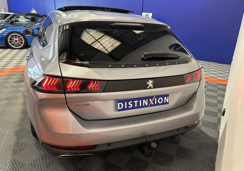 Vue 3/4 arrière d'une Peugeot 508 SW gris Platinium avec feux allumés et attelage visible en intérieur showroom.