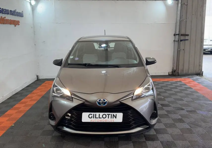 Vue avant d'une Toyota Yaris hybride gris foncé avec phares allumés dans un garage éclairé.