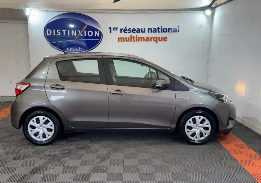 Vue latérale droite d'une Toyota Yaris hybride gris foncé, avec jantes blanches et badge Hybrid visible sur l'aile avant.
