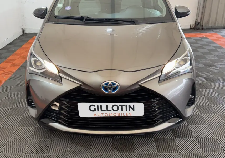 Vue avant rapprochée d’une Toyota Yaris hybride gris foncé avec phares allumés et calandre noire distinctive.