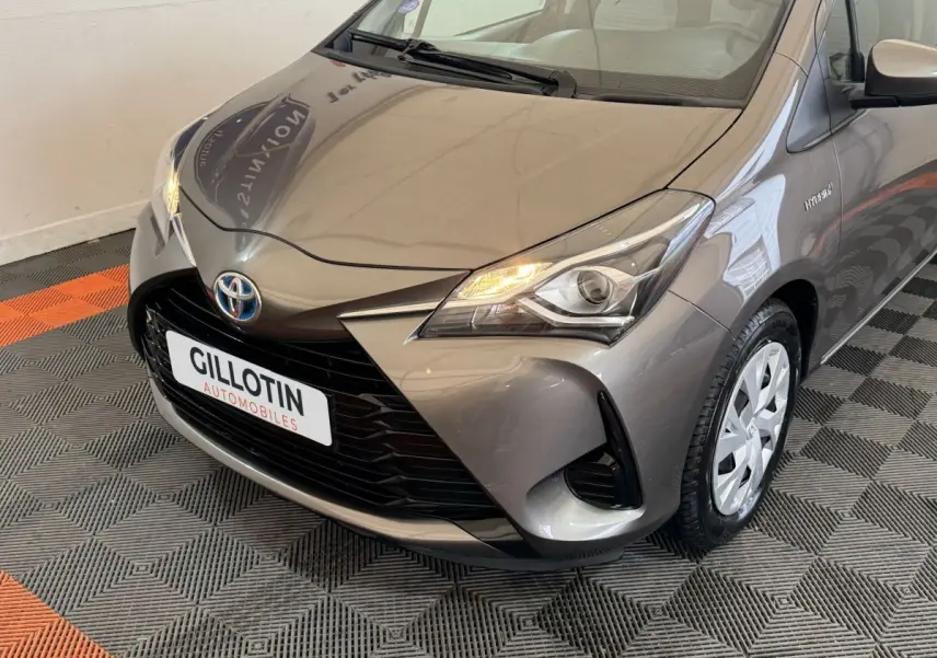 Vue 3/4 avant droit d'une Toyota Yaris hybride gris foncé avec phares allumés et jantes acier.