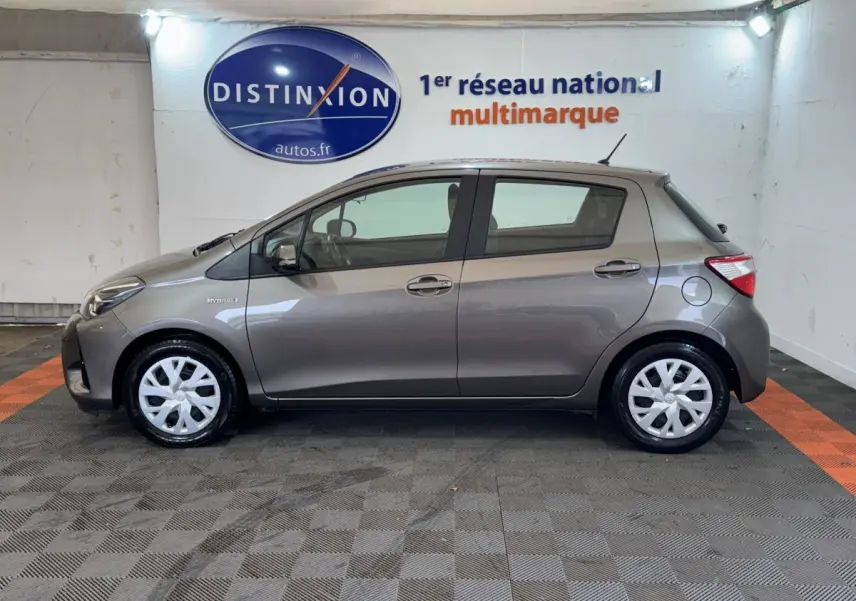 Vue de profil côté gauche d'une Toyota Yaris hybride gris foncé avec jantes blanches en intérieur showroom.