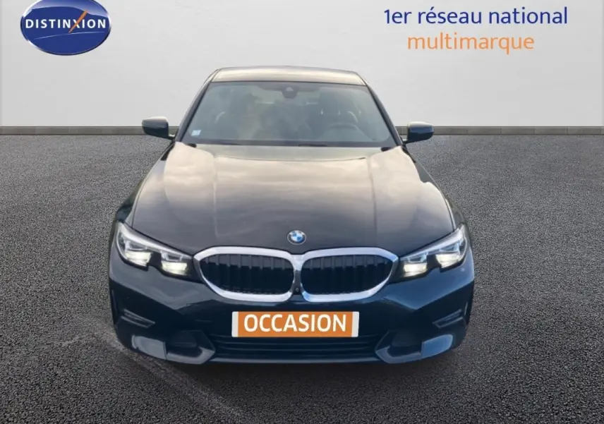 Vue avant d'une BMW Série 3 G20 noire de 2019 avec calandre chromée et phares allumés sur parking.