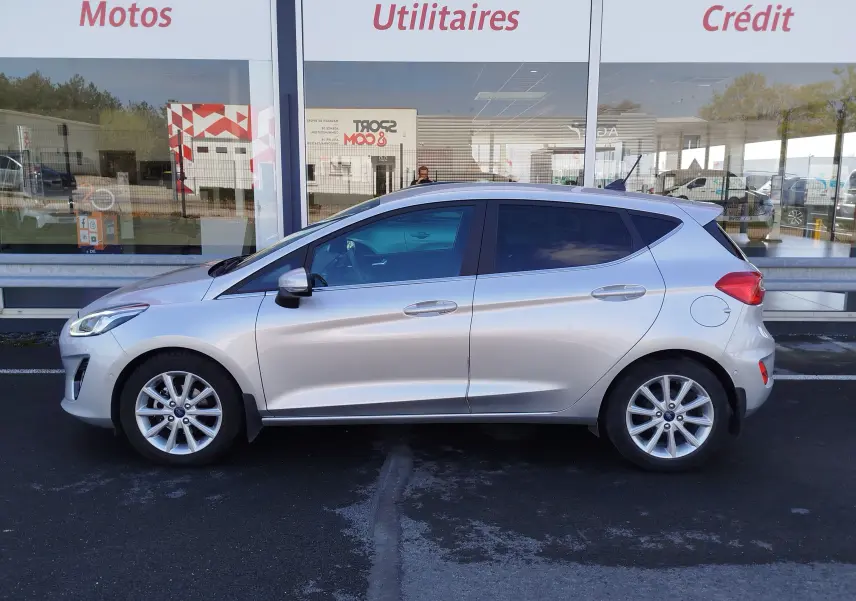 Profil côté gauche d'une Ford Fiesta gris lunaire 2019, finition Titanium, garée devant une vitrine commerciale.
