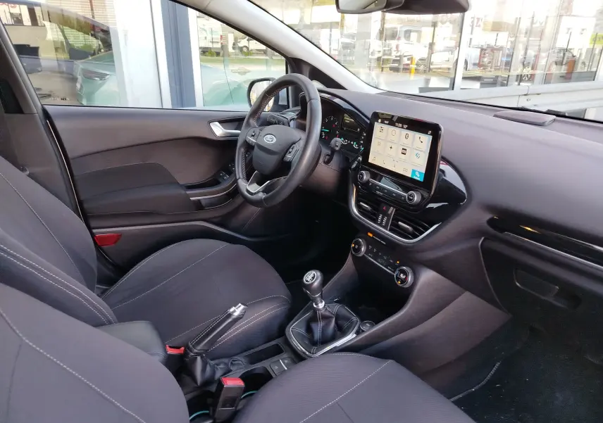 Vue intérieure côté conducteur de la Ford Fiesta 2019, tableau de bord avec écran tactile et sièges tissu gris foncé.