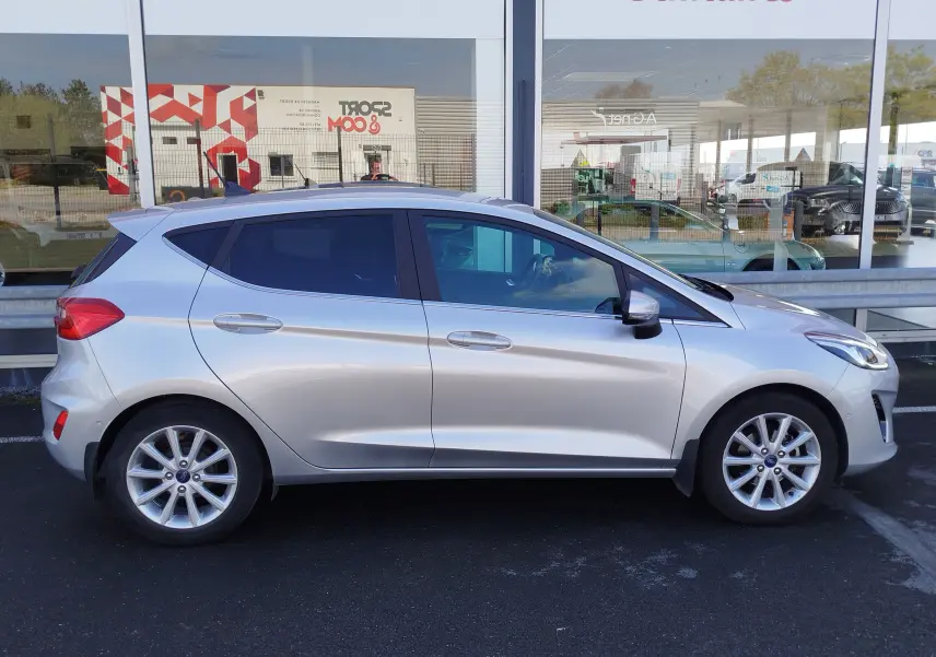 Vue de profil côté gauche d'une Ford Fiesta gris lunaire 2019 garée devant une vitrine, avec jantes alliage visibles.